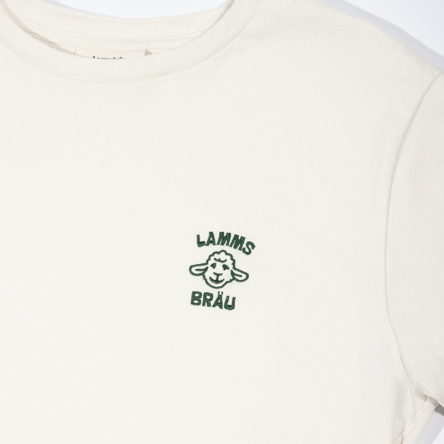 Weißes Kurzarm-T-Shirt in Vorderansicht-Detailaufnahme auf neutralem Hintergrund. In der Mitte der Brust ist ein grünes gesticktes Logo mit einem lächelnden Lamm-Kopf abgebildet, darüber der Text "LAMMS" und darunter "BRÄU" in grüner Schrift. Der gerippte Rundhalsausschnitt ist oben sichtbar.