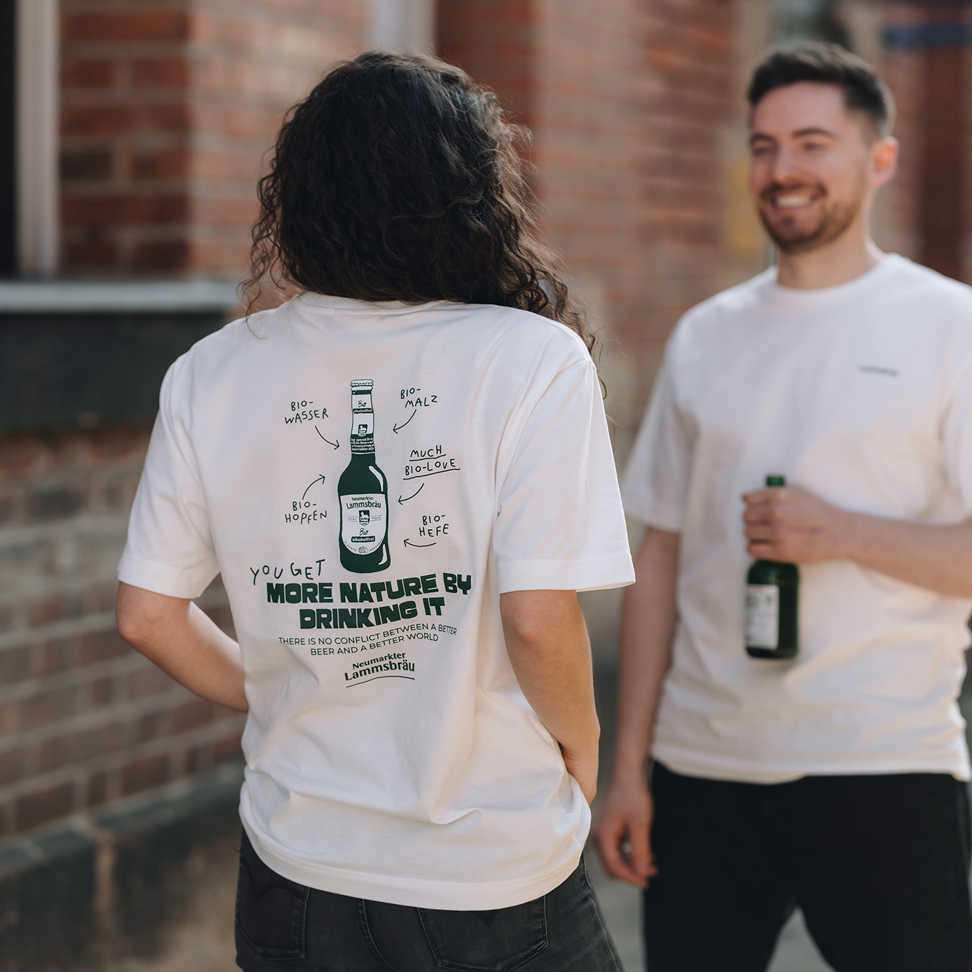 Das Bild zeigt den Rücken einer Frau und im Hintergrund einen Mann. Beide tragen ein weißes T-Shirt, auf dem Rücken der Frau sieht man den Rückenprint des T-Shirts: Illustration einer Bierflasche mit Zutatenlabeln (Bio-Wasser, Bio-Malz, Bio-Hopfen, Bio-Hefe) und Text „YOU GET MORE NATURE BY DRINKING IT"