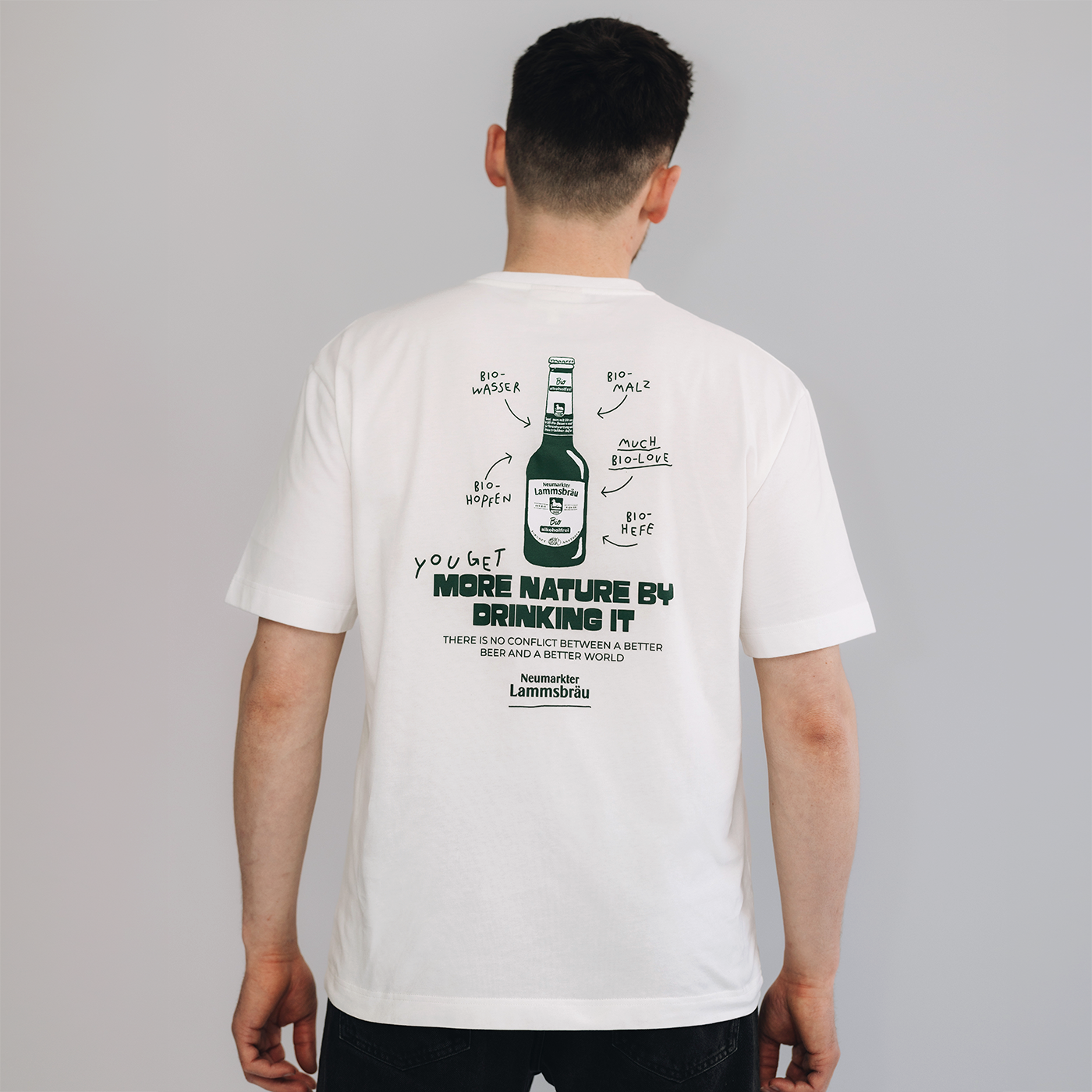 Auf dem Bild sieht man den Rücken eines Mannes. Der Mann trägt ein weißes T-Shirt mit Rückenprint: Bierflasche-Illustration mit Bio-Zutaten-Beschriftungen und Schriftzug „YOU GET MORE NATURE BY DRINKING IT". Der Mann ist vor einem neutralen grauen Hintergrund. 