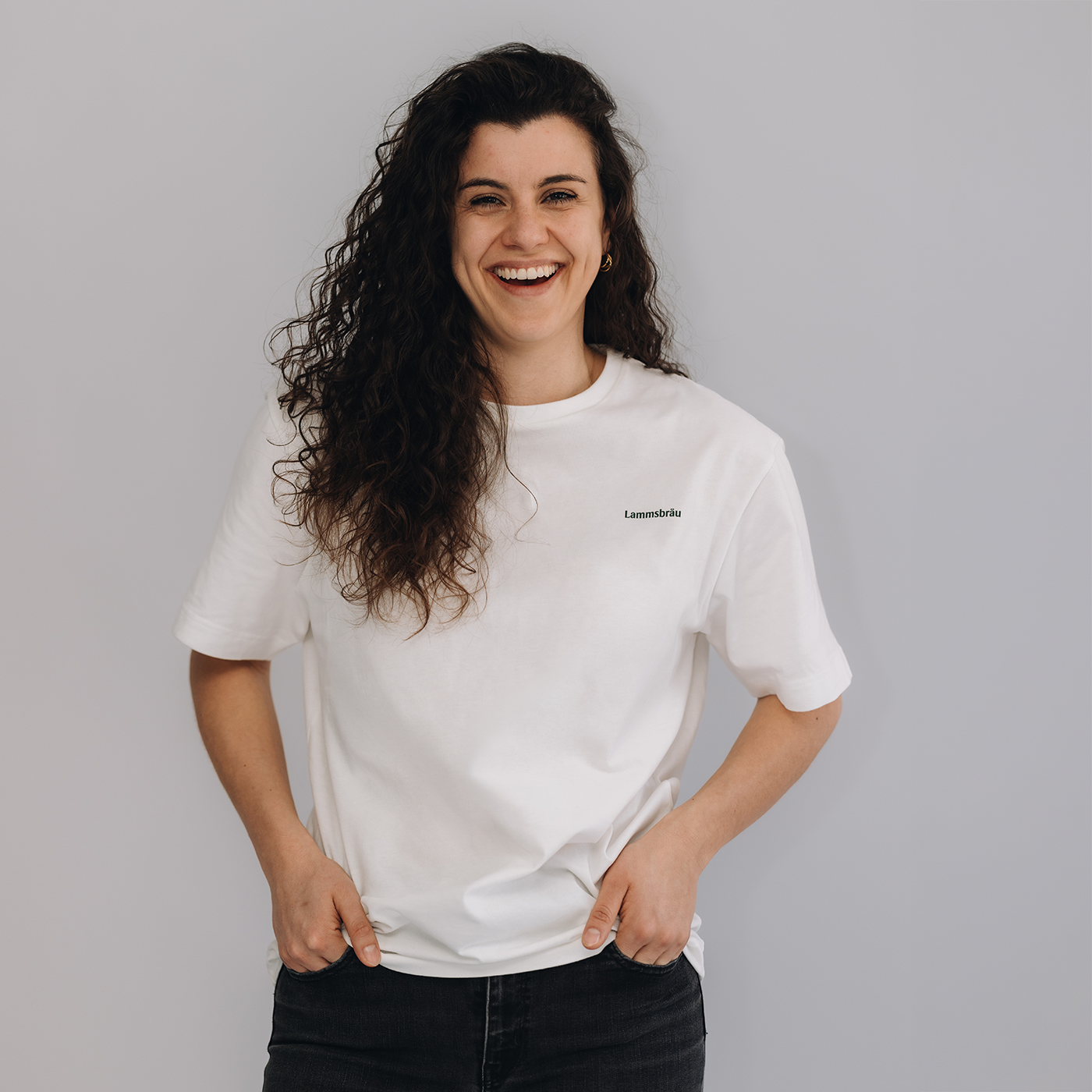 Auf dem Bild ist eine dunkelhaarige Frau vor einem neutralen weißen Hintergrund. Die Frau trägt ein weißes T-Shirt mit einem kleinen gedruckten Lammsbräu-Schriftzug auf Brusthöhe.
