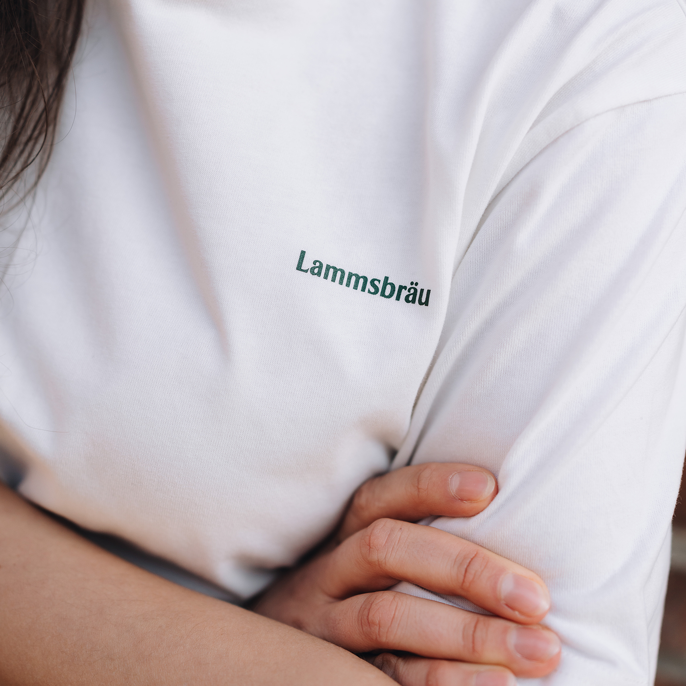 Nahaufnahme eines weißen T-Shirts getragen von einer Person. Der grüne Schriftzug "Lammsbräu" ist auf der Brustpartie in mittlerer Größe aufgedruckt. Im Vordergrund sichtbar sind die Hand und der Arm der Person.