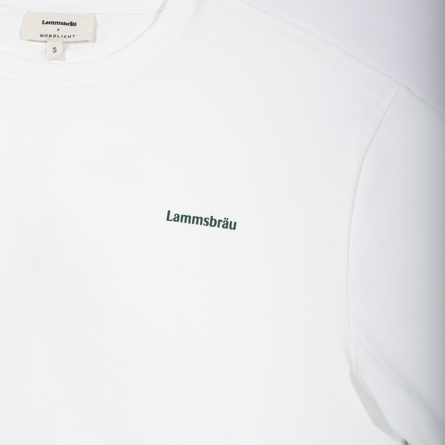 Weißes Kurzarm-T-Shirt in Vorderansicht auf neutralem Hintergrund. In der Mitte der Brust befindet sich der grüne Schriftzug "Lammsbräu". Im Kragen ist das beige Markenlabel und die Größenangabe.