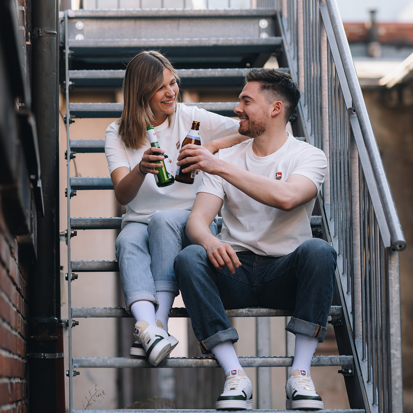 Zwei Personen sitzen auf einer Metalltreppe und stoßen mit zwei Bierflaschen an. Beide tragen weiße T-Shirts mit Lammsbräu-Logo.
