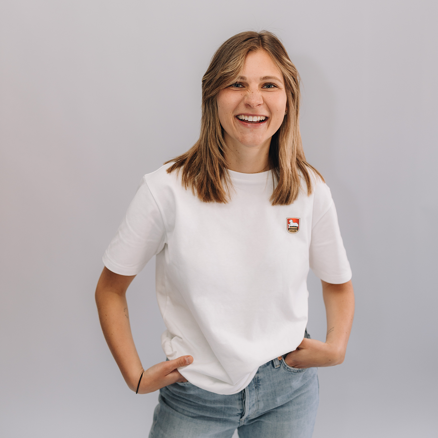 Frau trägt weißes T-Shirt mit kleinem aufgedruckten Lammsbräu-Wappen auf der Brust. Die Frau hat beide Hände in den Hosentaschen und steht vor einem neutralem grauen Hintergrund. 