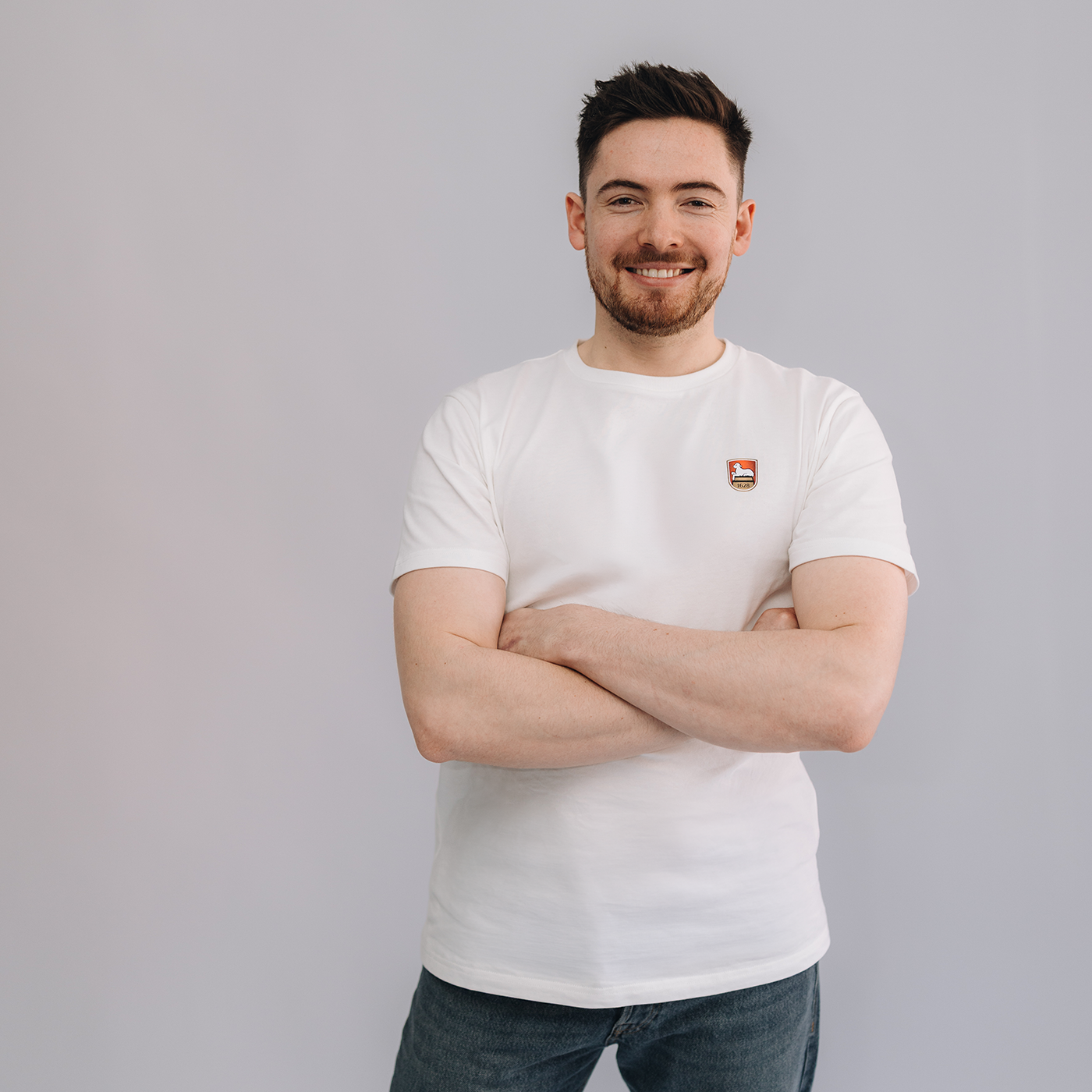 Mann trägt weißes T-Shirt mit aufgedrucktem Lammsbräu-Wappen auf der Brust. Der Mann hat die Arme verschränkt und steht vor einem  neutralen grauen Hintergrund.