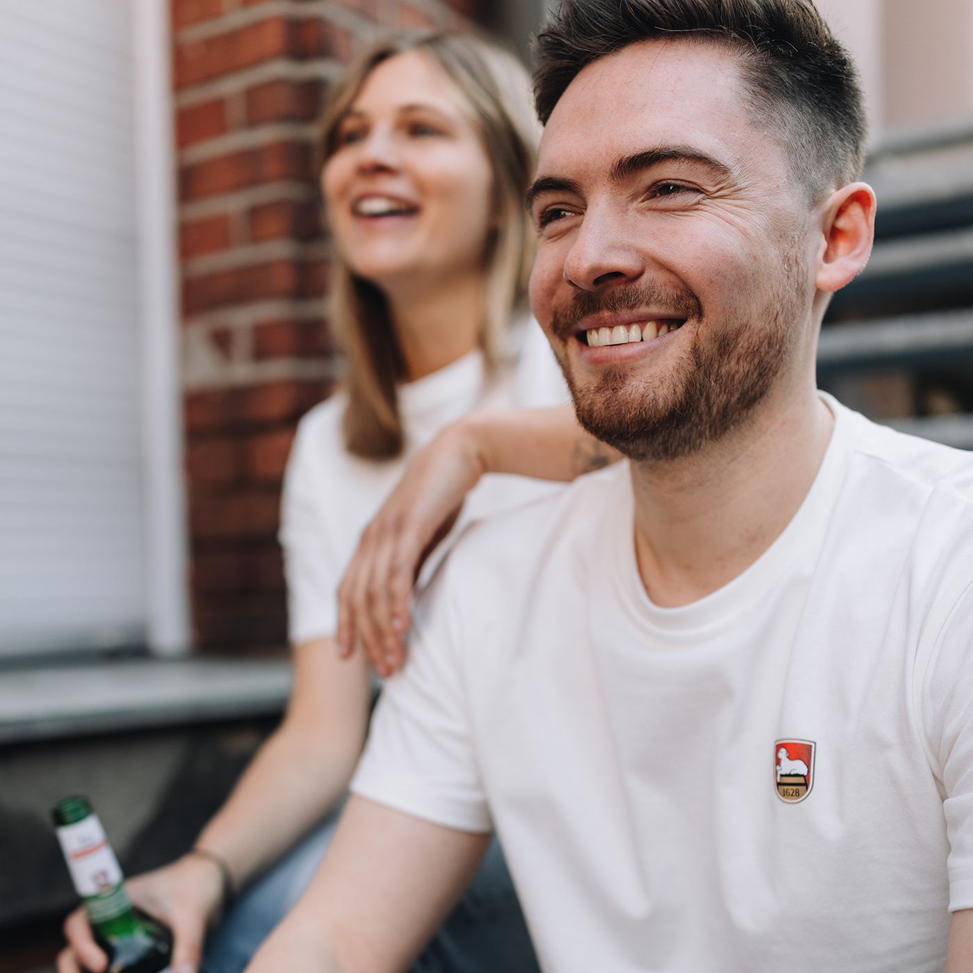 Mann und Frau sitzen nebeneinander auf einer Außentreppe vor einer roten Backsteinwand, beide tragen weiße T-Shirts mit einem kleinem aufgedruckten Lammsbräu-Wappen auf der Brust und halten Bierflaschen in den Händen.