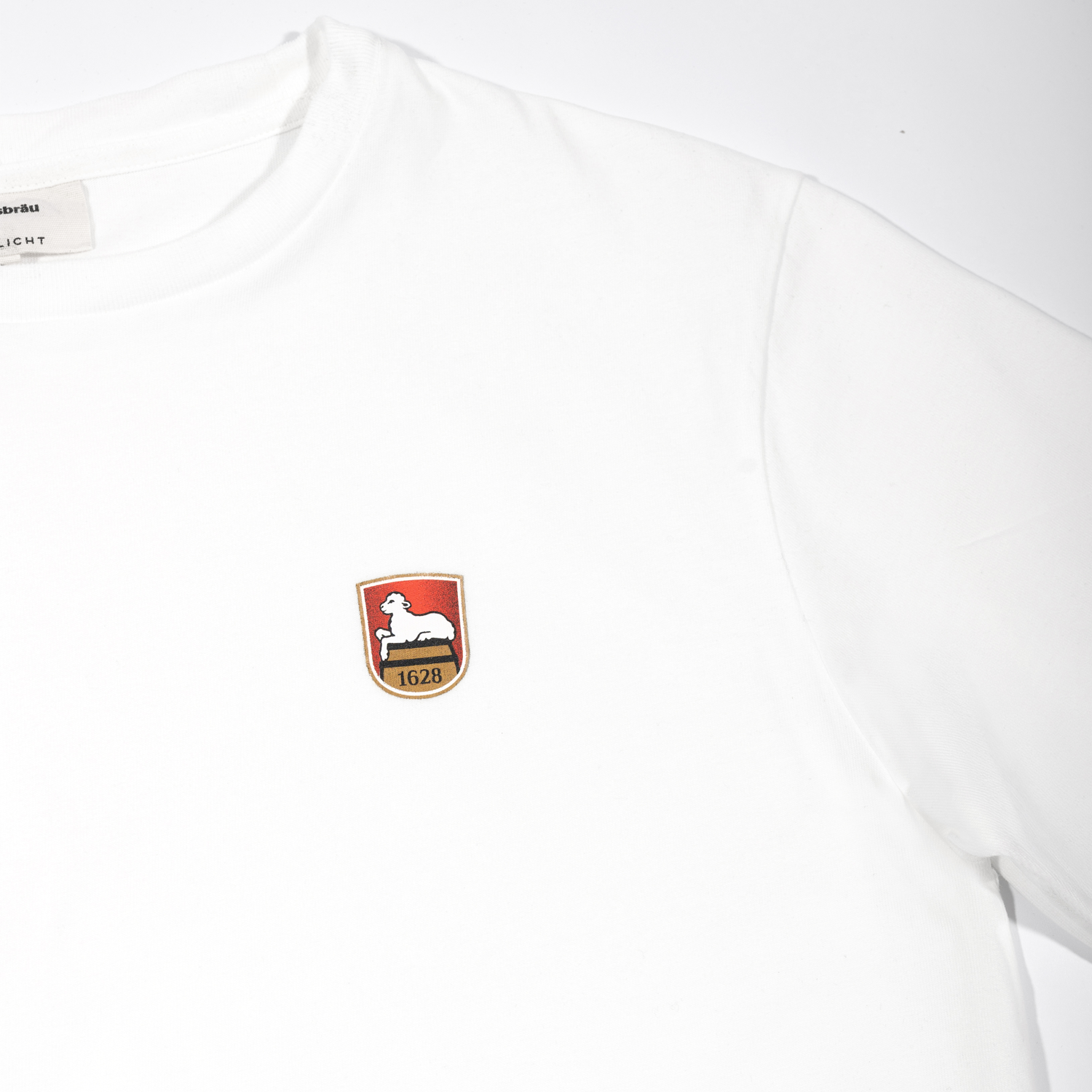 Detailansicht auf weißes Kurzarm-T-Shirt mit gedrucktem Lammsbräu-Wappen auf der Brust: Wappenschild mit weißem Lamm auf rotem Hintergrund, goldenem Fass Motiv und der Jahreszahl 1628.