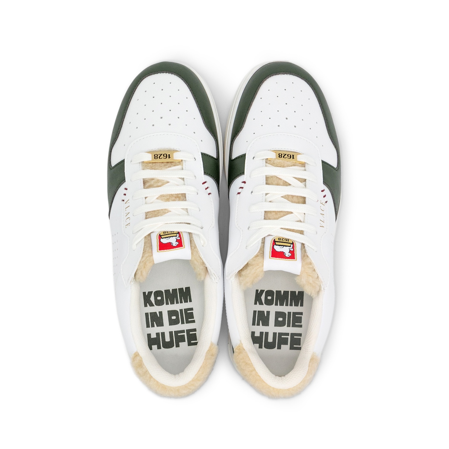 Das Bild zeigt ein Paar weiße Sneaker in Draufsicht auf weißem Hintergrund. Dunkelgrüne Seitenpaneele und cremefarbenes Futter sind sichtbar. Die Zungen zeigen die Markenlabels mit rot-weißem Wappenschild und goldenem "1628"-Label. Das beige Futter trägt auf beiden Innenseiten die Aufschrift "KOMM IN DIE HUFE".