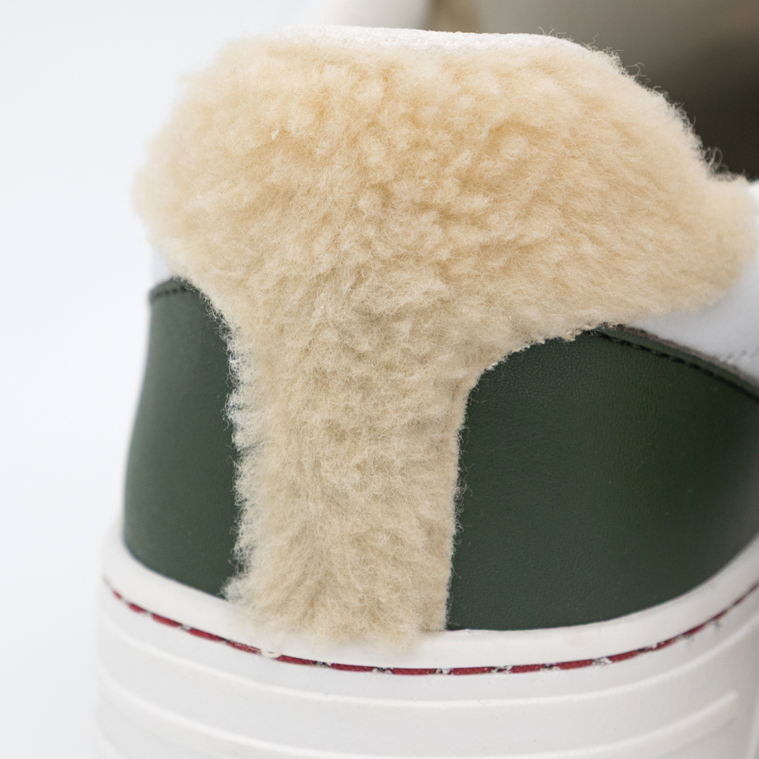 Nahaufnahme des Schaftbereichs eines weißen Sneakers mit flauschigem cremefarbenem Futter. Der Schuh zeigt dunkelgrüne Seitenpaneele und einen weißen Rand mit rot-goldener Nahtlinie. Das cremefarbene Fleece-Material erstreckt sich über den gesamten sichtbaren Schaftbereich.