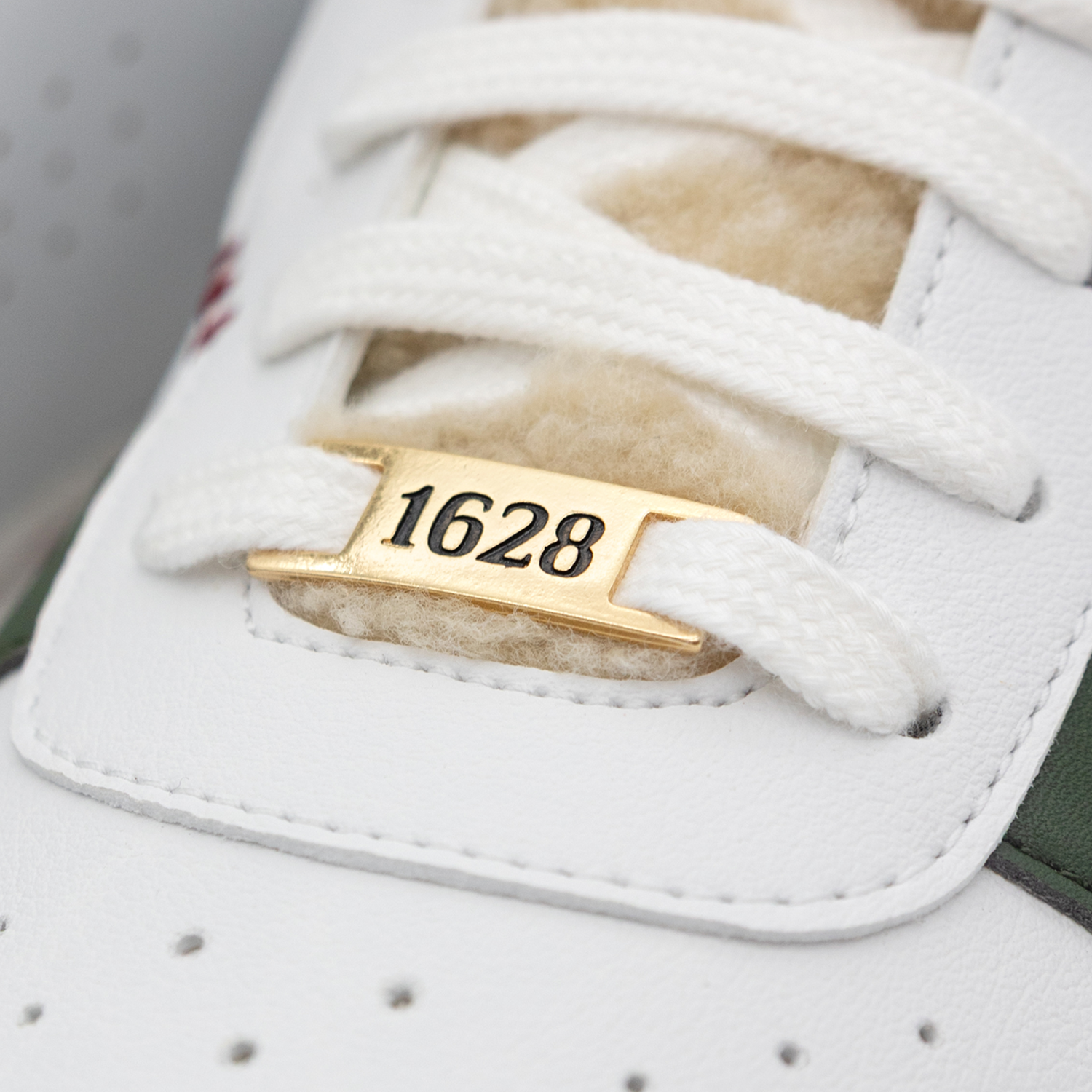 Detailaufnahme eines weißen Sneakers. Die weißen Schnürsenkel verlaufen diagonal über cremefarbenes Fleece-Futter. Dazwischen ein goldenes metallisches Label mit schwarzer Aufschrift "1628". 
