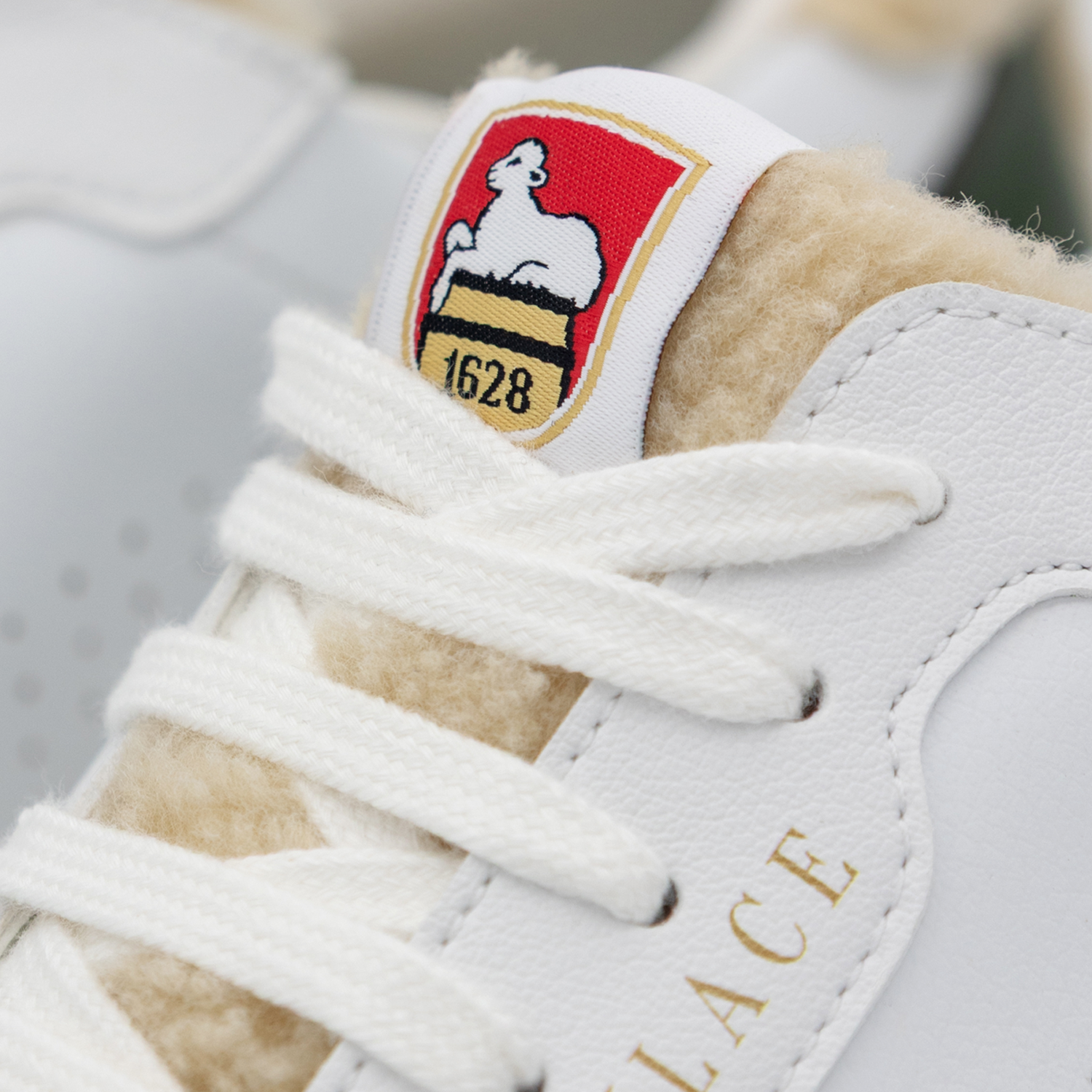 Detailaufnahme eines weißen Sneakers. Ein weißes Zungen-Label trägt das farbig gestickte Neumarkter Lammsbräu-Wappen: weißes Lamm auf rotem Grund, goldbegrenzter Fassboden mit schwarzen Streifen und schwarzer Jahreszahl "1628" darunter. Weiße Schnürsenkel und cremefarbenes Futter sind sichtbar.