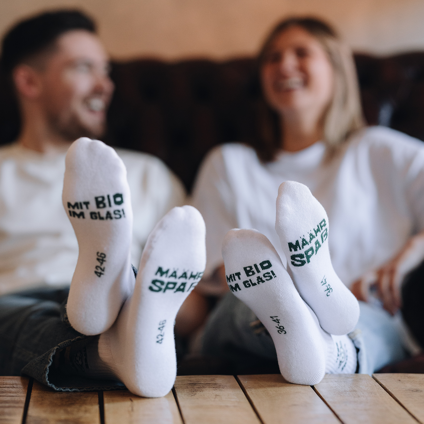 Das Bild zeigt zwei Paar weiße Socken mit Webung in grüner Farbe auf den Sohlen. Im Hintergrund sitzen eine Frau und ein Mann in hellen T-Shirts auf einem dunklen Sofa, die beiden Personen tragen die Socken. 