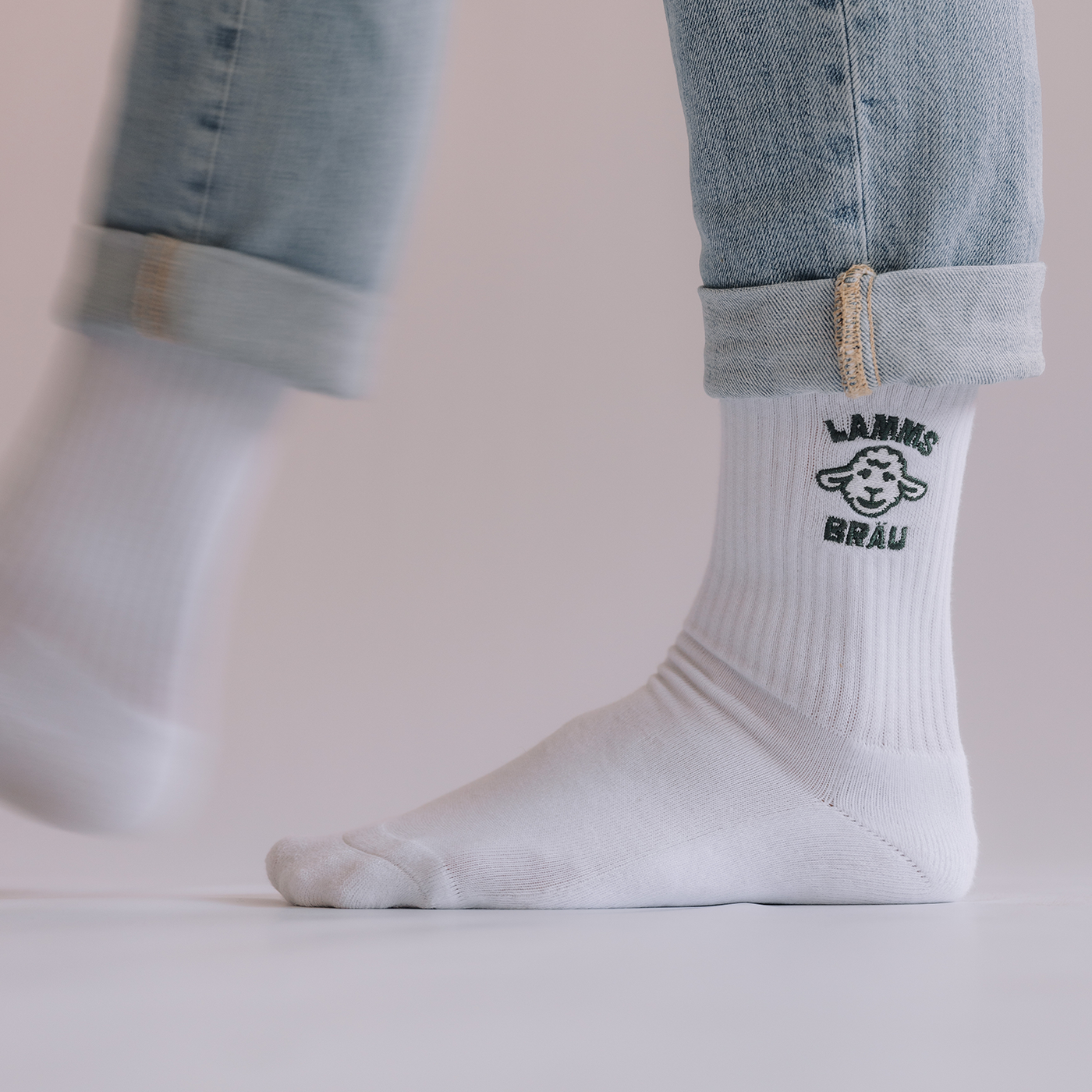 Nahaufnahme eines weißen Socken mit dunkelgrünen Lamm-Kopf-Motiv und Schriftzug auf der Außenseite, getragen unter einer hochgekrempelten hellblauen Jeans.