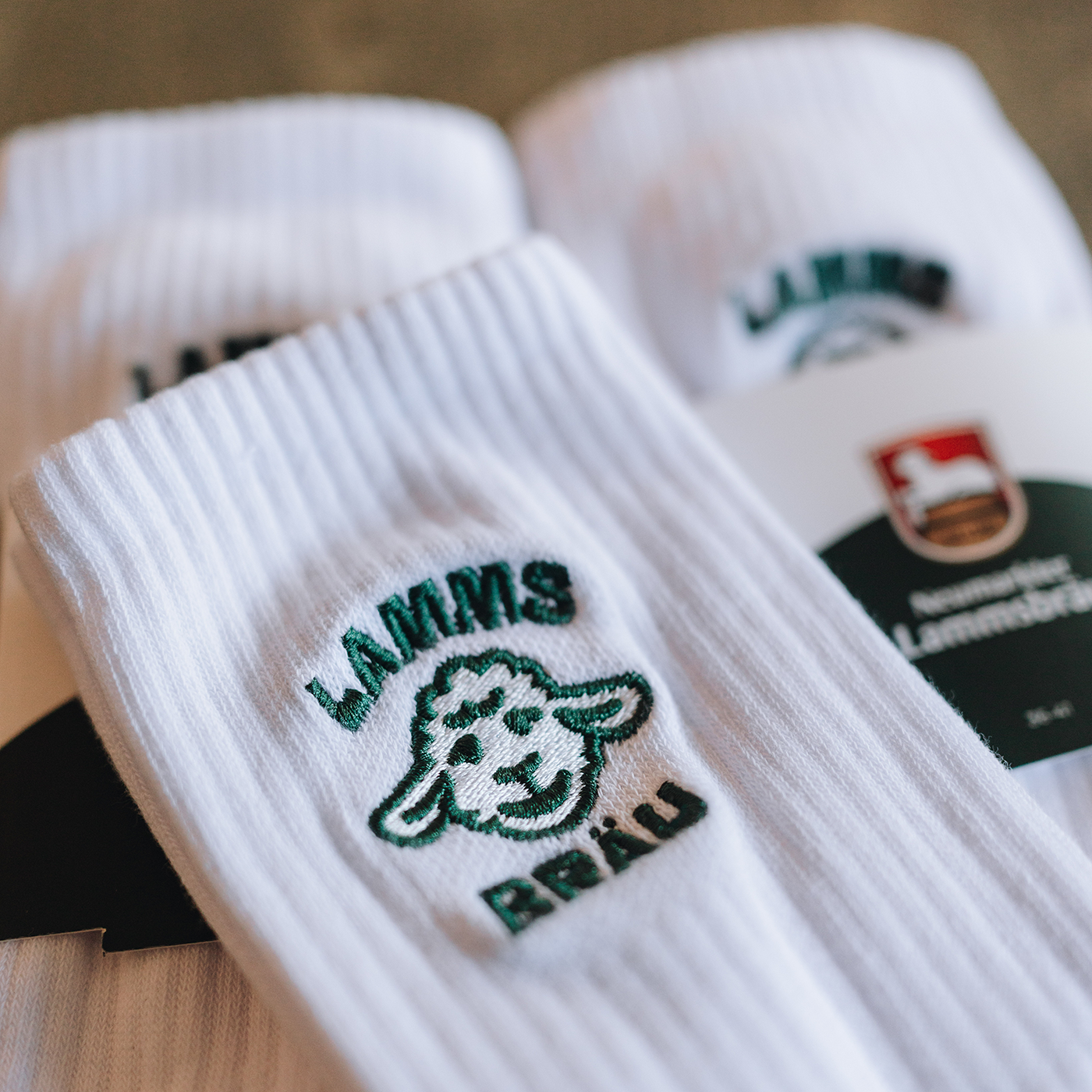 Weiße Socken mit dunkelgrünem Lamm-Kopf-Motiv und Schriftzug, liegend auf einer Oberfläche neben ihrer Produktverpackung mit schwarz-grünem Design.