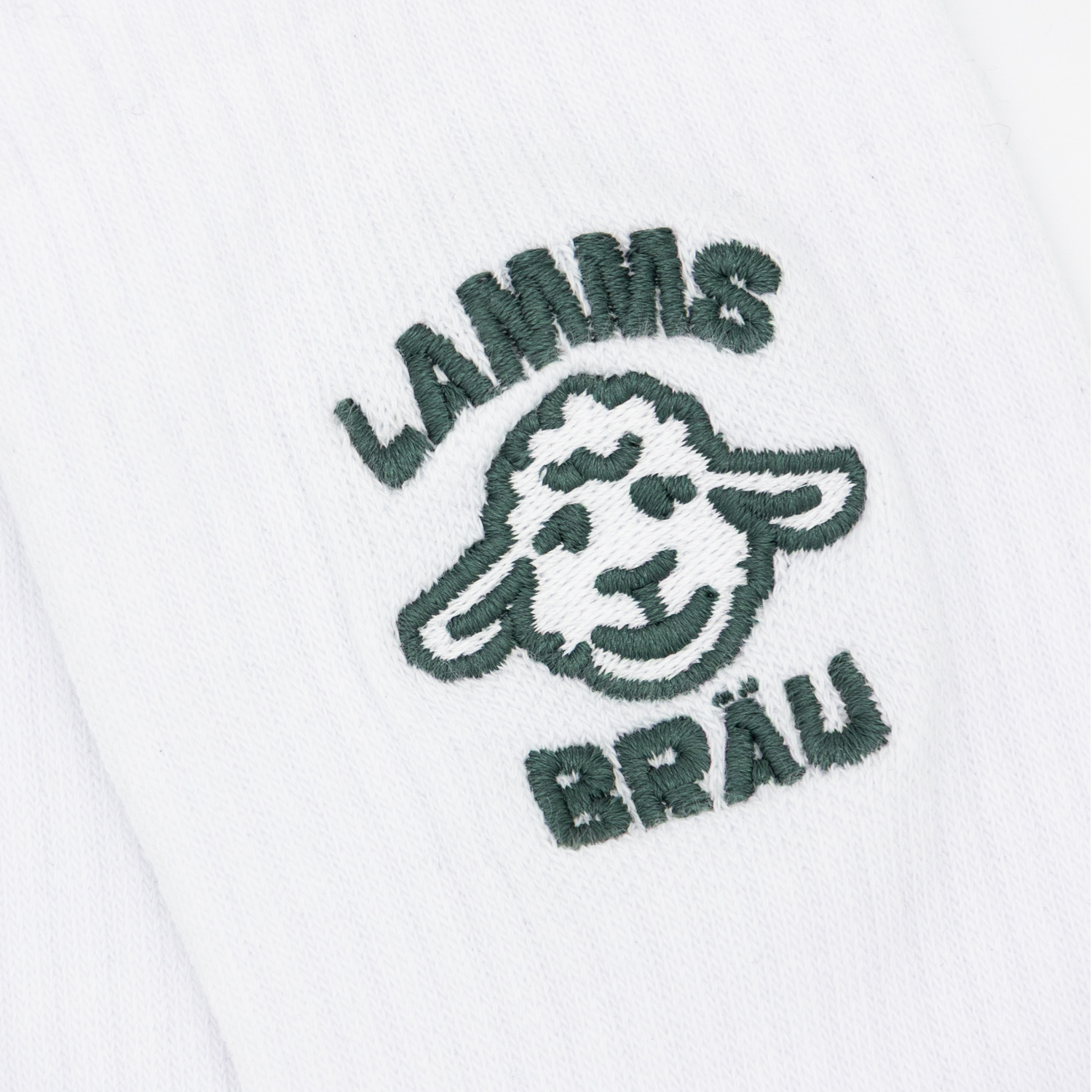Detailaufnahme einer weißen Socke mit gesticktem Motiv in Dunkelgrün: stilisierter Lamm-Kopf mit der Aufschrift "LAMMS" oben und "BRÄU" unten, auf neutralem Hintergrund.