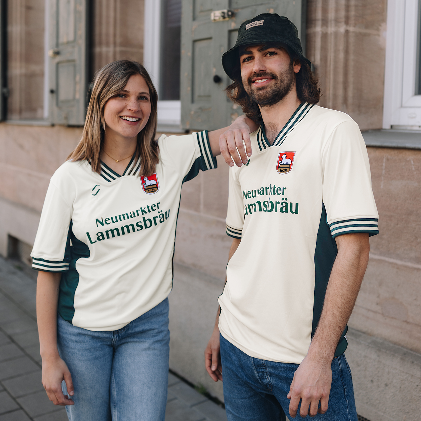 Ein weibliches Model und ein männliches Model stehen nebeneinander. Das weibliche Model stützt sich mit einem Arm am männlichen Model ab. Beide Models tragenein Retro-Fußballtrikot. Das Trikot ist im Farbton "elfenbein" und hat auf der Brust den Schriftzug "Neumarkter Lammsbräu". Außerdem sind das Logo von runamics und das Logo von Lammsbräu auf dem Trikot integriert. Das männliche Model trägt noch einen dunkelgrünen Fischerhut mit Lammsbräu-Patch.