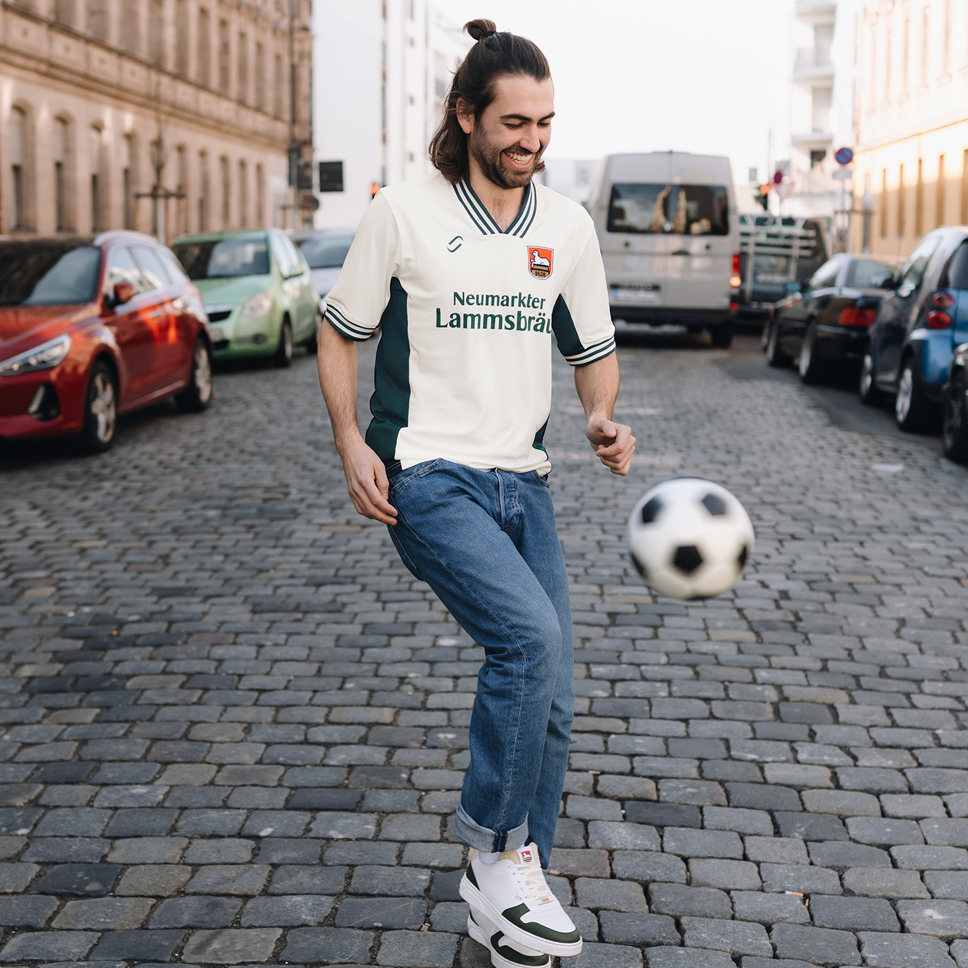 Ein männliches Model trägt ein Retro-Fußballtrikot. Das Trikot ist im Farbton "elfenbein" und hat auf der Brust den Schriftzug "Neumarkter Lammsbräu". Außerdem sind das Logo von runamics und das Logo von Lammsbräu auf dem Trikot integriert. Das Model spielt mit einem Fußball.