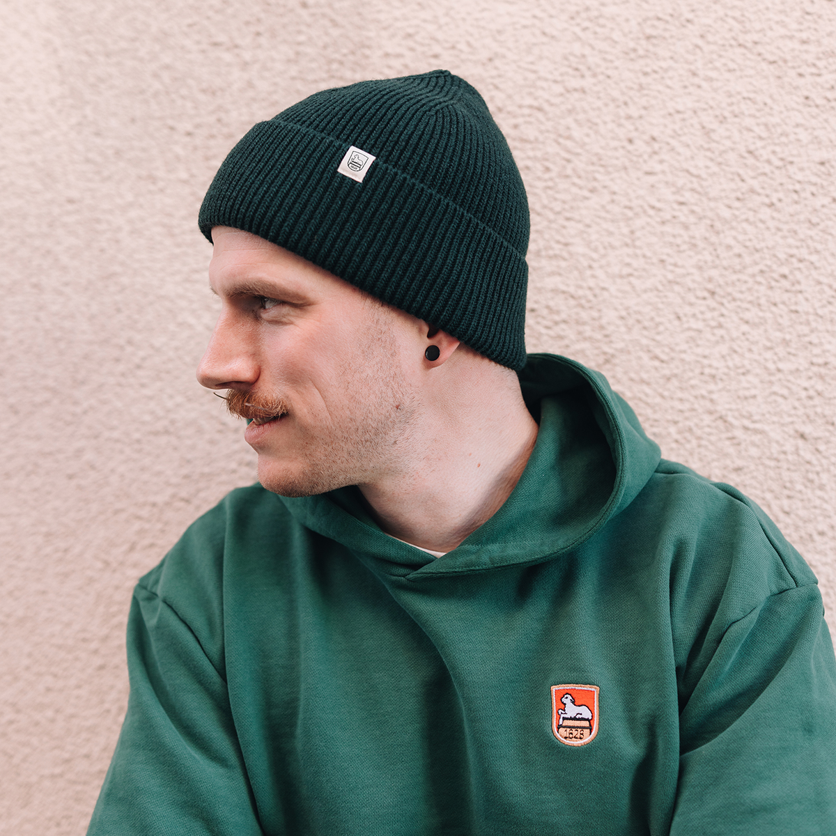 Seitenansicht eines Mannes, der eine dunkelgrüne Mütze mit seitlich platziertem, hellem Logo-Patch trägt. Er ist zusätzlich mit einem farblich passenden grünen Hoodie gekleidet, der ein Wappen-Logo auf der Brust zeigt.