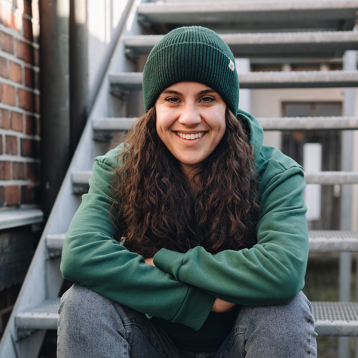 Frontalaufnahme einer lächelnden Frau, die auf einer Metalltreppe sitzt und eine dunkelgrüne Mütze mit Umschlag trägt. Das Outfit wird durch einen farblich abgestimmten grünen Kapuzenpullover ergänzt, wobei das helle Mützen-Etikett leicht seitlich sichtbar ist.
