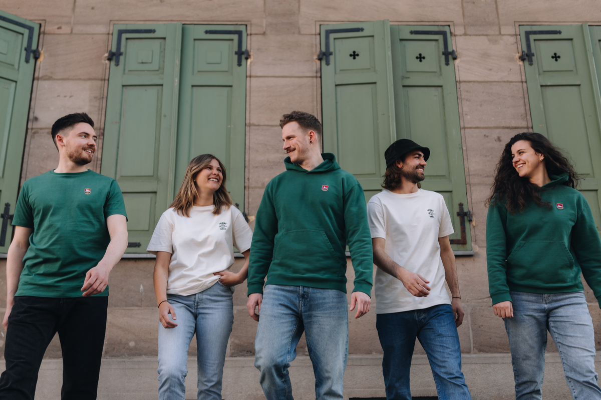 Auf dem Bild sind 5 Personen nebeneinander zu sehen, die sich gegenseitig ansehen. Sie tragen alle Lammsbräu-Merch. Es handelt es sich um drei Männer und zwei Frauen. Im Hintergrund ist die Fassade eines Gebäudes zu sehen.