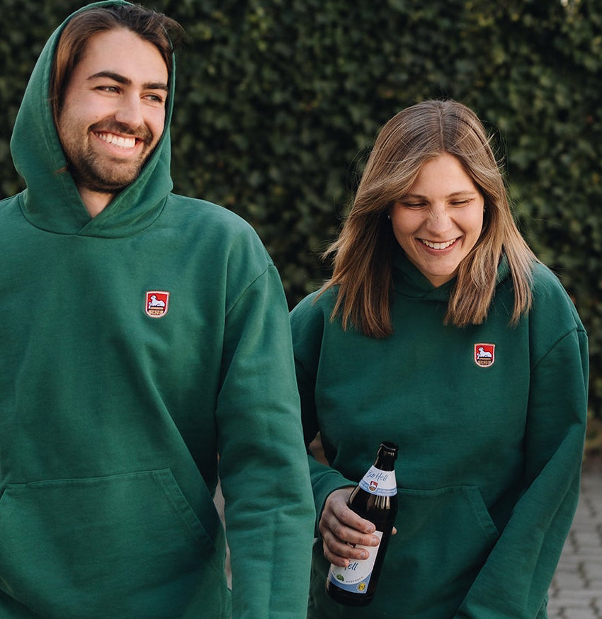 Eine Frau und ein Mann tragen dunkelgrüne Kapuzenpullover mit kleinem rotem Wappenstickerei-Logo auf der Brust. Die Frau hält eine dunkle Bierflasche mit weißem und blauem Etikett in der Hand. Beide lächeln in die Kamera und stehen vor einer unscharfen Efeu-Kulisse im Freien.