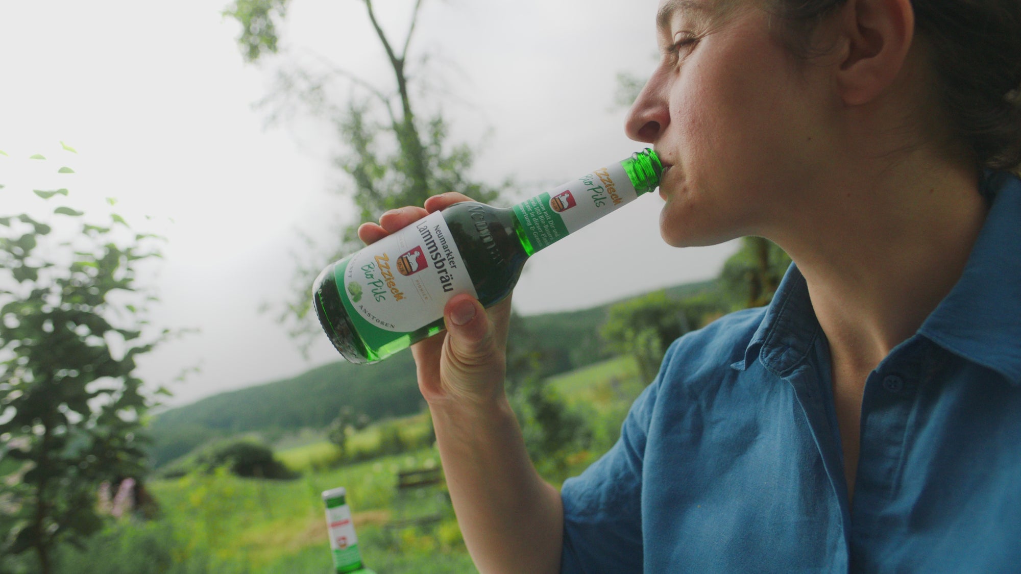 Eine Frau in blauem Oberteil trinkt aus einer grünen Bierflasche mit weiß-grünem Etikett von Neumarkter Lammsbräu. Die Szene ist in seitlicher Nahaufnahme vor unscharfem Hintergrund mit Bäumen und Hügellandschaft im Freien aufgenommen.
